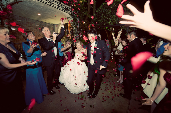 the happy couple leaving the reception while family and guests throw dark pink rose petals at them - bride is wearing a white ball gown style dress and - photo by Houston based wedding photographer Adam Nyholt 